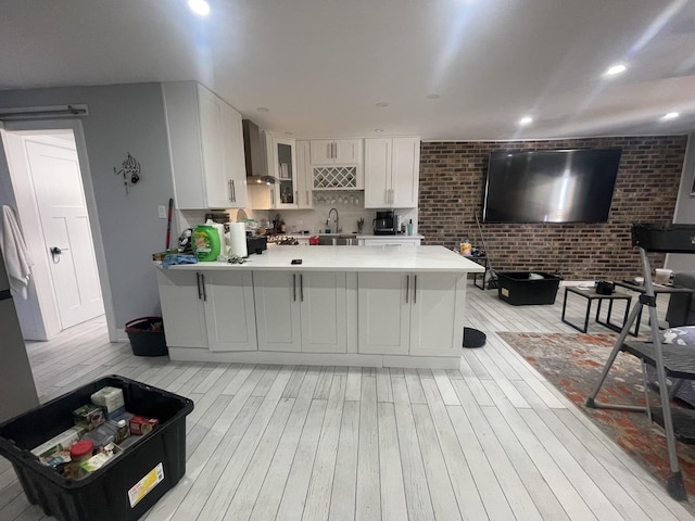 kitchen with a peninsula, a sink, wall chimney range hood, glass insert cabinets, and light countertops