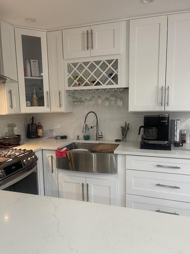 kitchen with a sink, gas range oven, white cabinetry, glass insert cabinets, and wall chimney exhaust hood