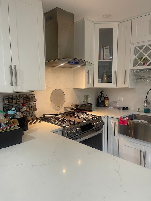 kitchen with light stone counters, stainless steel gas range, sink, white cabinets, and wall chimney exhaust hood