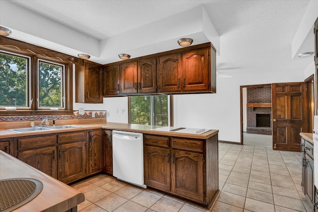 kitchen with white dishwasher, sink, kitchen peninsula, light tile patterned floors, and a brick fireplace