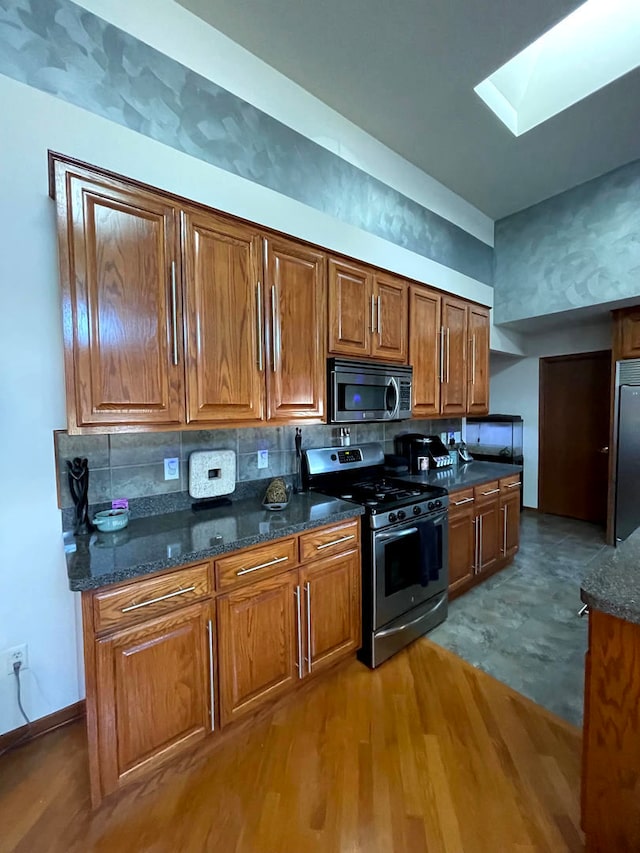 kitchen with a skylight, dark stone counters, stainless steel appliances, light wood-type flooring, and decorative backsplash