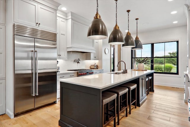 kitchen with decorative light fixtures, white cabinetry, sink, stainless steel built in fridge, and a kitchen island with sink