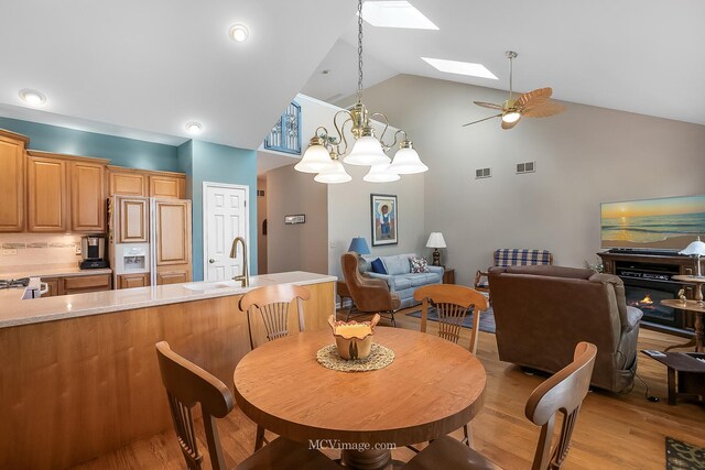 dining space featuring sink, light wood-type flooring, ceiling fan with notable chandelier, a skylight, and high vaulted ceiling