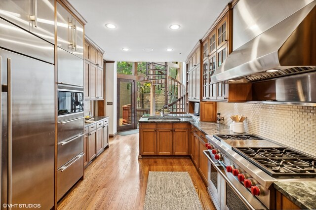 kitchen featuring high end appliances, stone countertops, wall chimney exhaust hood, backsplash, and light hardwood / wood-style floors