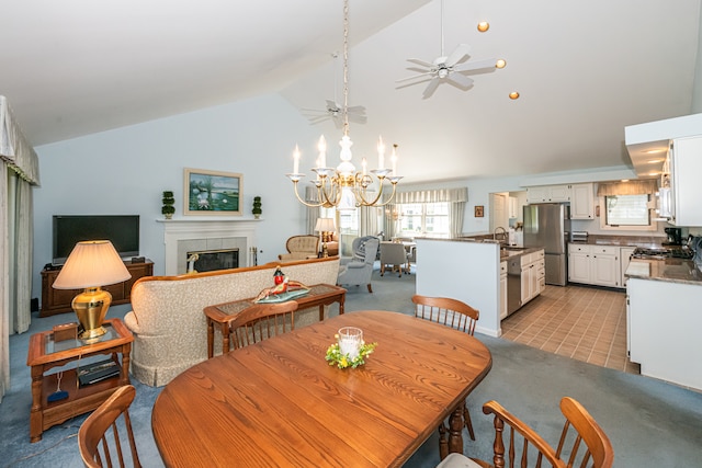 dining room with sink, high vaulted ceiling, a tiled fireplace, ceiling fan with notable chandelier, and light colored carpet