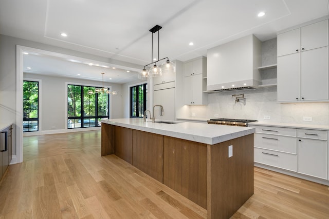 kitchen featuring an island with sink, light wood-type flooring, custom range hood, and white cabinetry