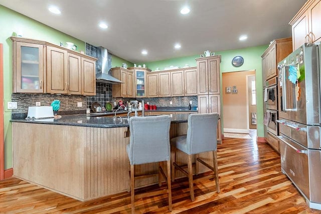 kitchen featuring light wood-type flooring, appliances with stainless steel finishes, backsplash, wall chimney exhaust hood, and kitchen peninsula