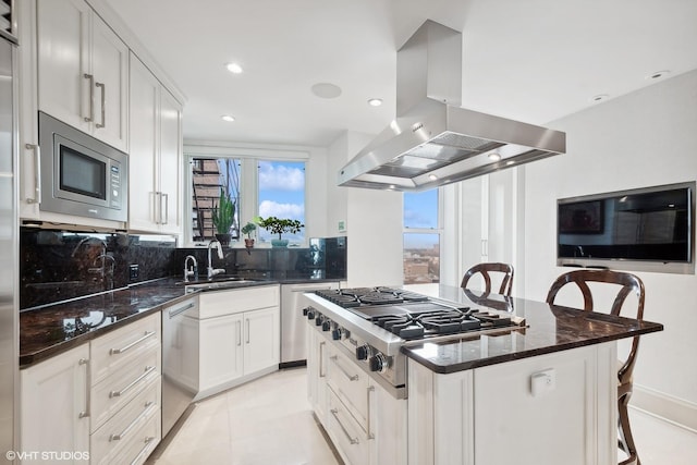 kitchen featuring appliances with stainless steel finishes, island range hood, white cabinets, a kitchen bar, and dark stone counters