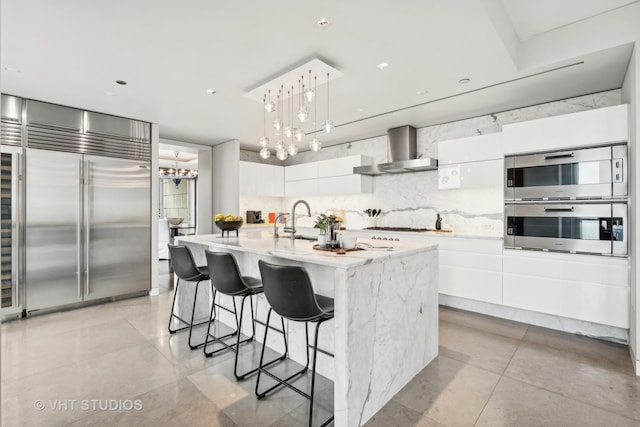 kitchen with decorative light fixtures, backsplash, appliances with stainless steel finishes, white cabinets, and wall chimney exhaust hood