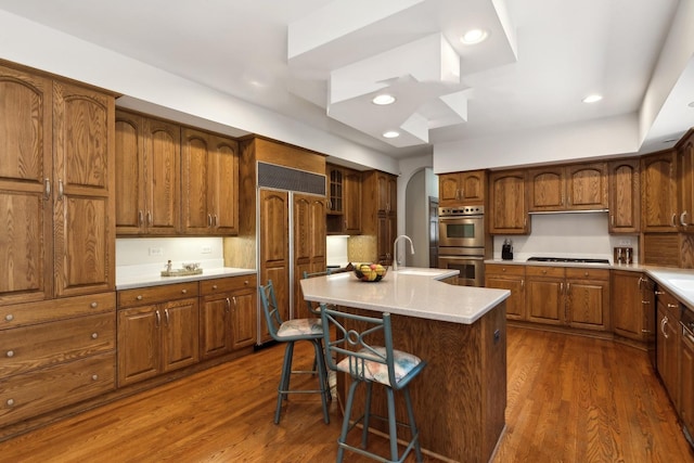 kitchen featuring brown cabinetry, a kitchen breakfast bar, a kitchen island with sink, light countertops, and stainless steel double oven