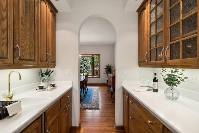 kitchen with arched walkways, light countertops, glass insert cabinets, dark wood-type flooring, and a sink