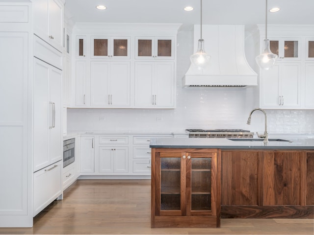 kitchen with hanging light fixtures, custom range hood, backsplash, light wood-type flooring, and sink