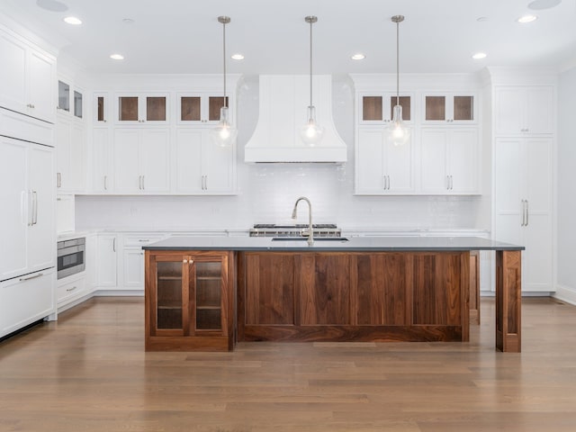 kitchen with a center island with sink, paneled fridge, hanging light fixtures, custom exhaust hood, and hardwood / wood-style floors