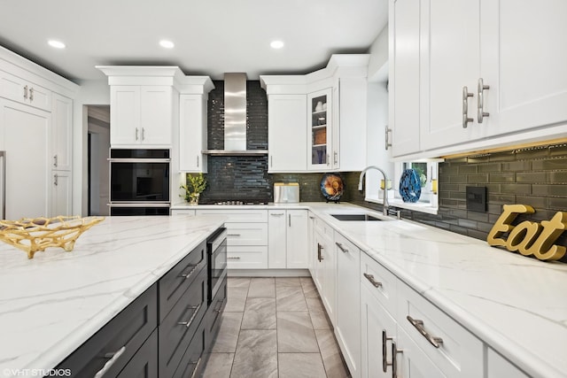 kitchen featuring wall chimney range hood, sink, light stone counters, black appliances, and white cabinets