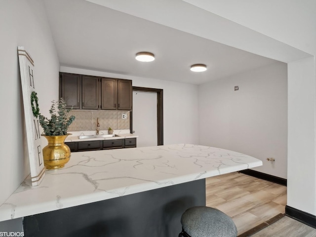 kitchen with light stone counters, backsplash, a sink, light wood-style flooring, and dark brown cabinets