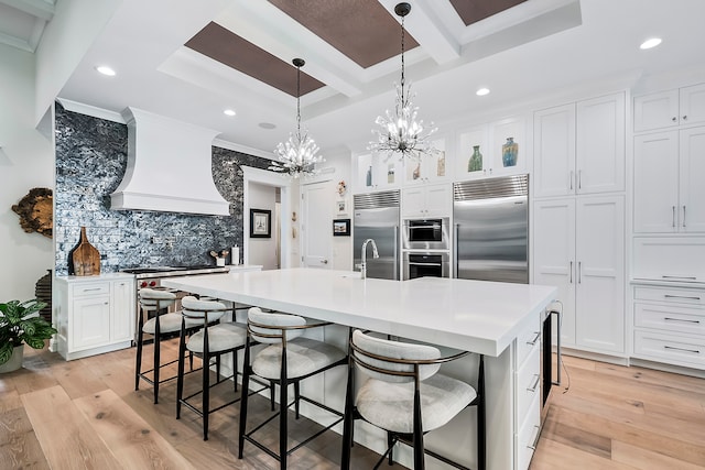 kitchen featuring stainless steel appliances, a chandelier, an island with sink, light wood-type flooring, and custom exhaust hood