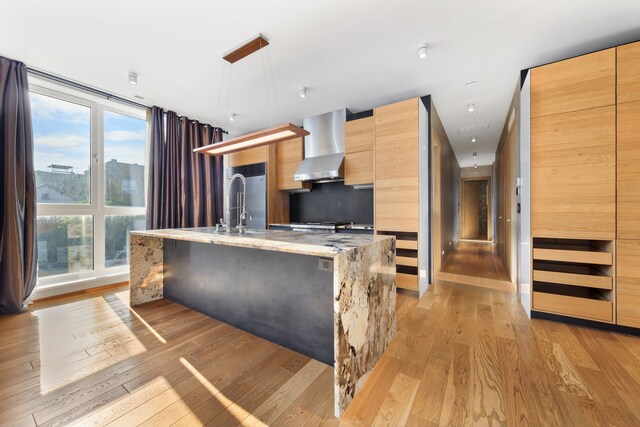 kitchen with wall chimney exhaust hood, light wood-type flooring, light stone countertops, pendant lighting, and a kitchen island with sink