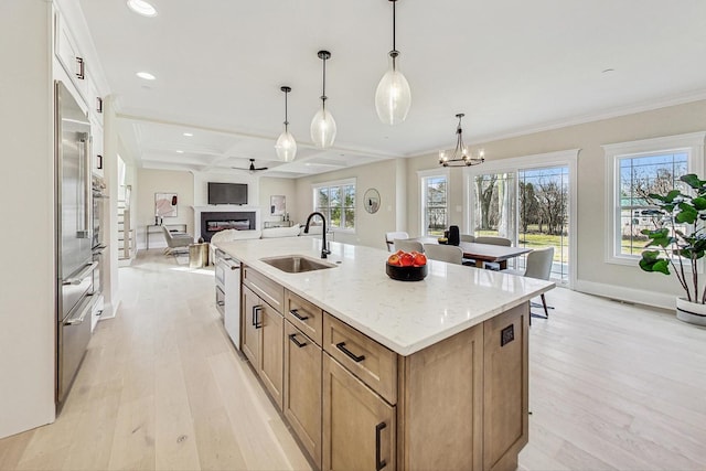 kitchen featuring a fireplace, light wood finished floors, a kitchen island with sink, a sink, and coffered ceiling