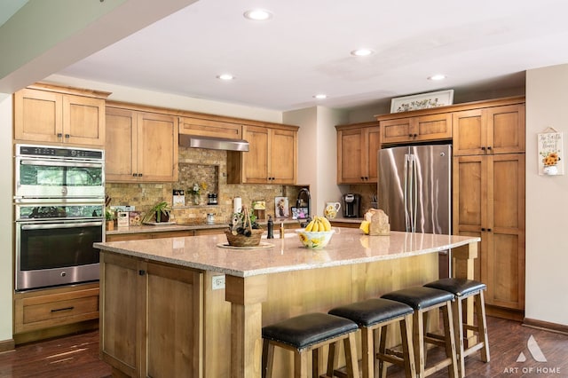 kitchen with wall chimney range hood, dark wood-type flooring, an island with sink, and stainless steel appliances