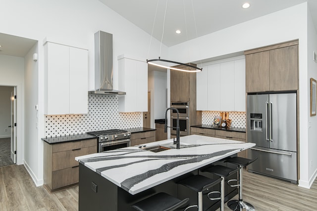 kitchen with backsplash, light hardwood / wood-style floors, a kitchen island with sink, wall chimney range hood, and premium appliances