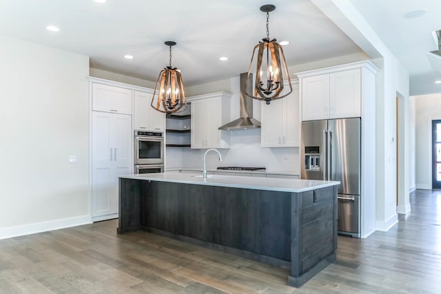 kitchen with a center island with sink, stainless steel appliances, light countertops, wall chimney range hood, and white cabinetry