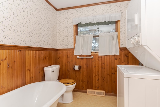 bathroom featuring wooden walls, toilet, stacked washing maching and dryer, a tub, and ornamental molding