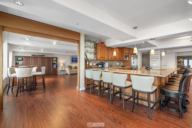 kitchen featuring brown cabinetry, dark wood-style flooring, decorative backsplash, appliances with stainless steel finishes, and a kitchen bar