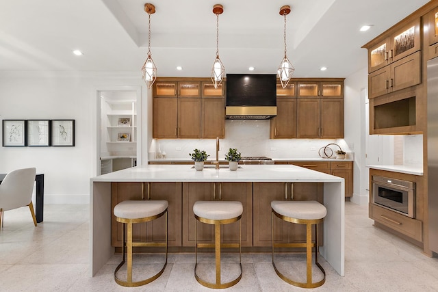 kitchen with baseboards, an island with sink, brown cabinets, premium range hood, and backsplash