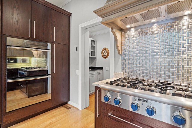 kitchen with light hardwood / wood-style flooring, dark brown cabinetry, tasteful backsplash, custom exhaust hood, and crown molding