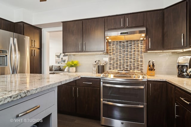 kitchen with appliances with stainless steel finishes, decorative backsplash, dark brown cabinets, light stone countertops, and range hood