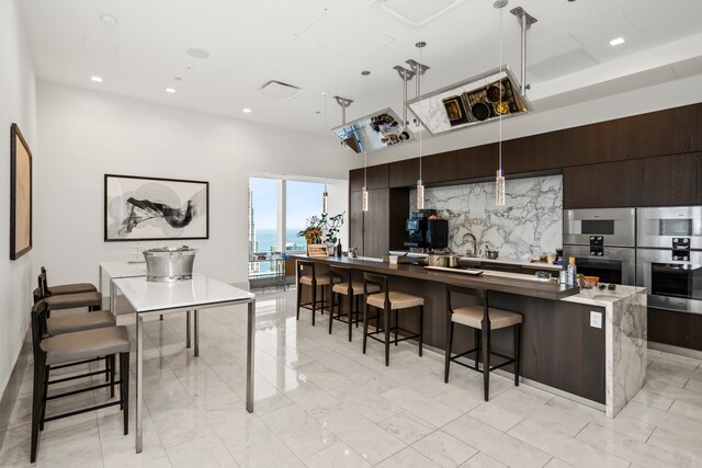 kitchen with dark brown cabinets, pendant lighting, an island with sink, decorative backsplash, and double oven