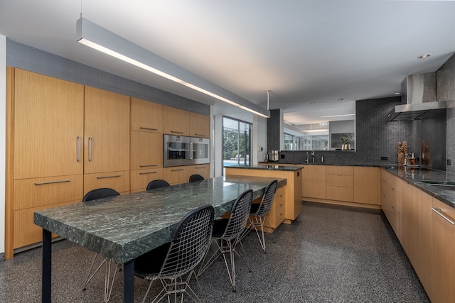 kitchen featuring stainless steel microwave, a kitchen bar, wall chimney range hood, decorative backsplash, and light brown cabinetry