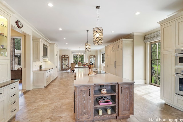 kitchen featuring a kitchen island with sink, pendant lighting, cream cabinetry, crown molding, and an inviting chandelier