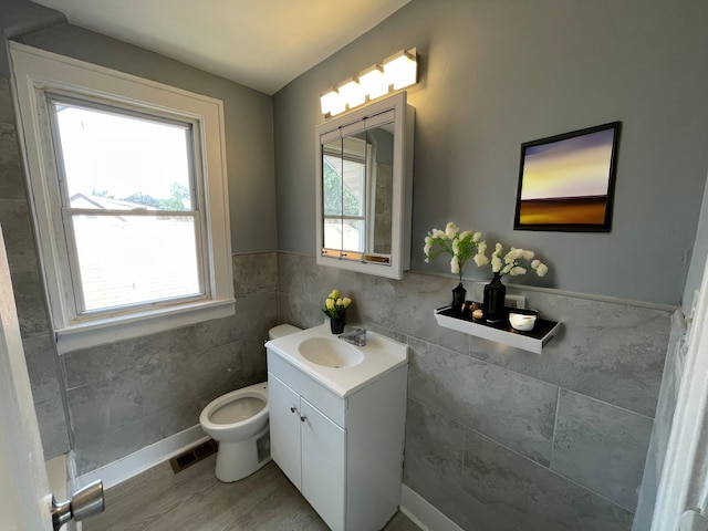 bathroom with toilet, plenty of natural light, tile walls, wood-type flooring, and vanity