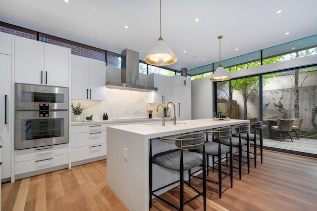 kitchen with light wood-type flooring, stainless steel double oven, decorative light fixtures, a center island with sink, and wall chimney range hood