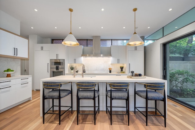 kitchen with light wood-type flooring, white cabinetry, wall chimney range hood, an island with sink, and decorative backsplash