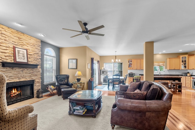 living room featuring sink, a stone fireplace, ceiling fan with notable chandelier, and light wood-type flooring