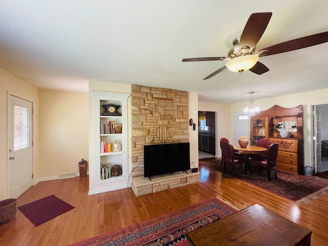 living room featuring ceiling fan with notable chandelier, built in features, wood-type flooring, and a stone fireplace