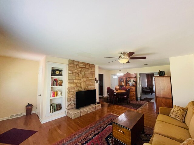 living room featuring dark wood-type flooring, built in features, ceiling fan, and a stone fireplace