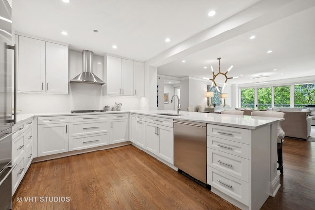 kitchen with a sink, open floor plan, stainless steel appliances, a peninsula, and wall chimney range hood