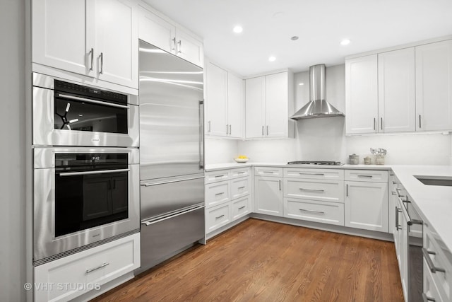 kitchen with light countertops, stainless steel appliances, wood finished floors, white cabinetry, and wall chimney exhaust hood