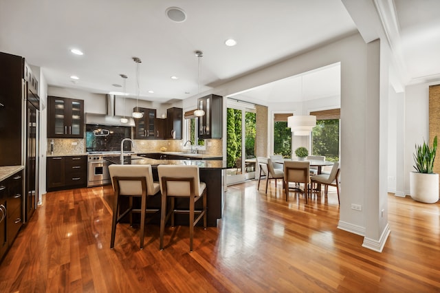 kitchen with dark wood-type flooring, a kitchen bar, dark brown cabinetry, a kitchen island with sink, and wall chimney range hood