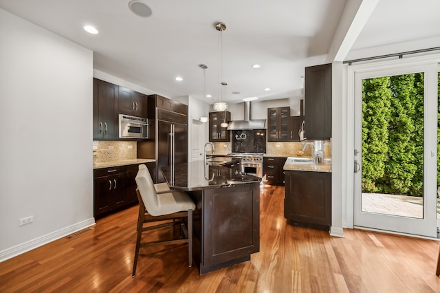 kitchen with light hardwood / wood-style floors, high end appliances, wall chimney range hood, and an island with sink