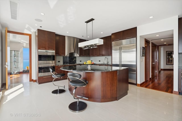 kitchen featuring decorative light fixtures, a center island, dark stone countertops, stainless steel appliances, and wall chimney range hood