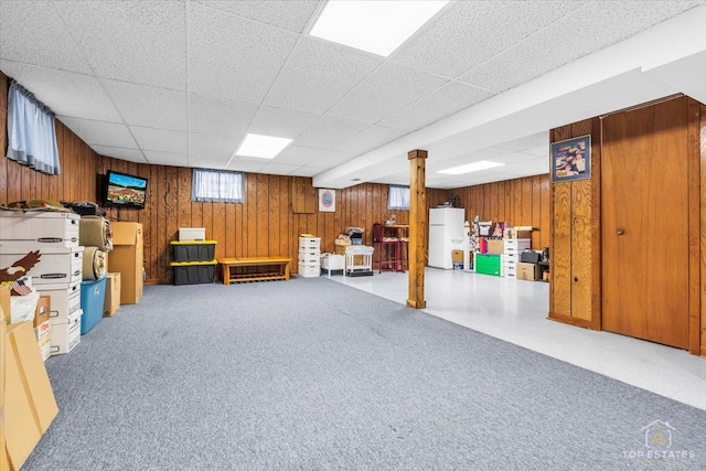 basement with a paneled ceiling, wood walls, carpet flooring, and freestanding refrigerator