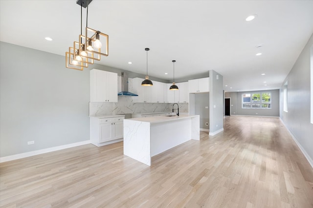 kitchen with decorative light fixtures, wall chimney exhaust hood, light hardwood / wood-style flooring, backsplash, and a kitchen island with sink