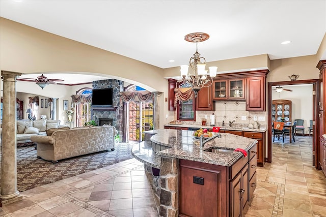 kitchen with a kitchen island with sink, ceiling fan with notable chandelier, decorative columns, sink, and a stone fireplace