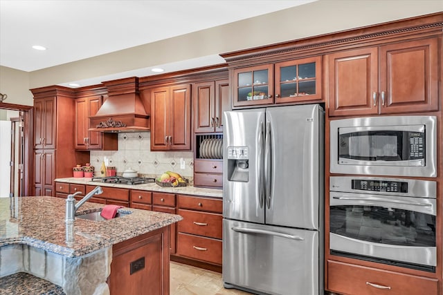 kitchen featuring light stone countertops, custom range hood, appliances with stainless steel finishes, sink, and decorative backsplash