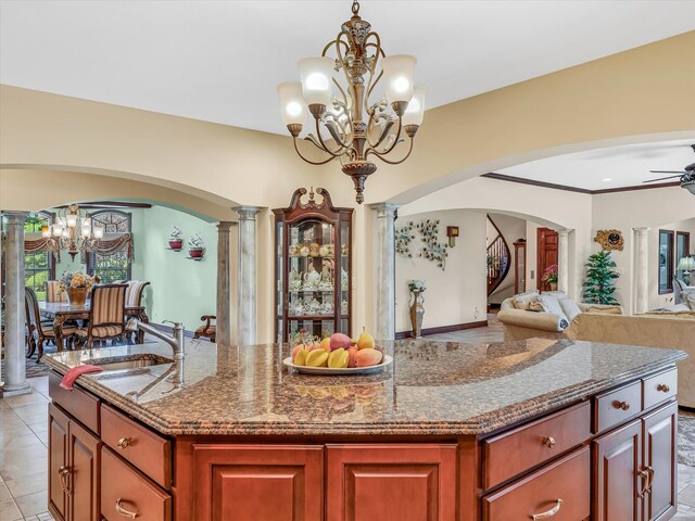 kitchen with stone counters, ceiling fan with notable chandelier, decorative columns, sink, and an island with sink