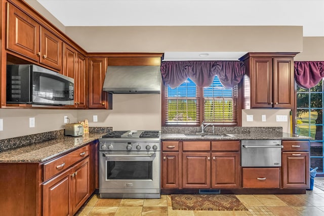 kitchen with dark stone countertops, appliances with stainless steel finishes, sink, and wall chimney range hood